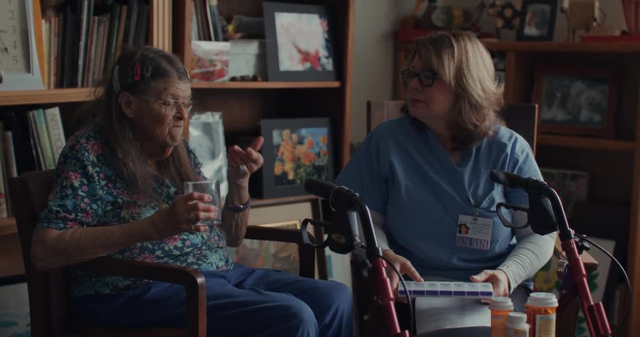 Two women are sitting in a room one of them holds a transparent cup while the other carries a notepad and wears medical scrubs