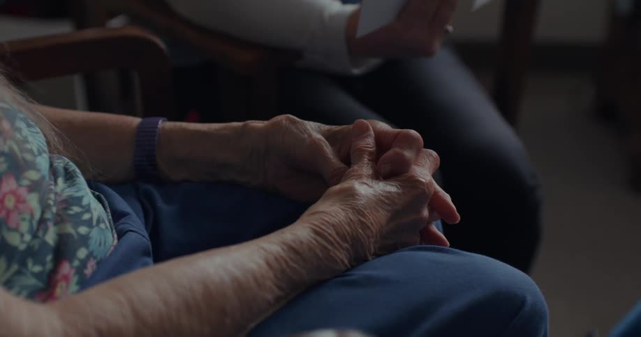 A woman sits with crossed hands as another woman holds pieces of paper