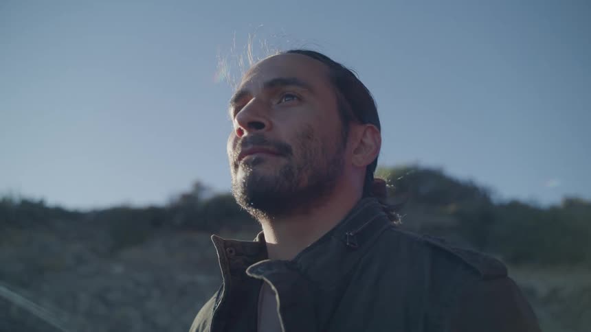 A bearded man stands outdoors looking at the sky on a sunny day