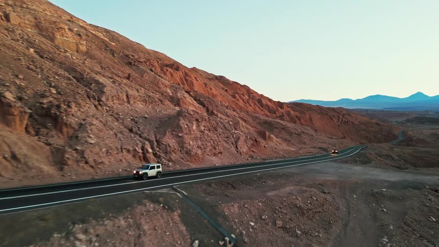 Two cars drive down a paved road surrounded by rocky mountains