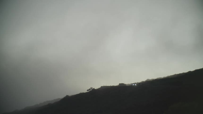 A car with headlights on drives on a hilly terrain under a cloudy sky