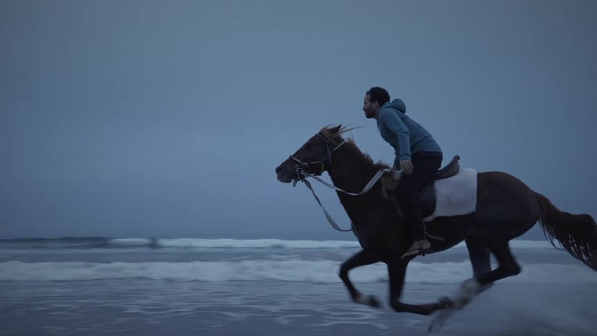 A man galloping on a horse along the sea line on a beach