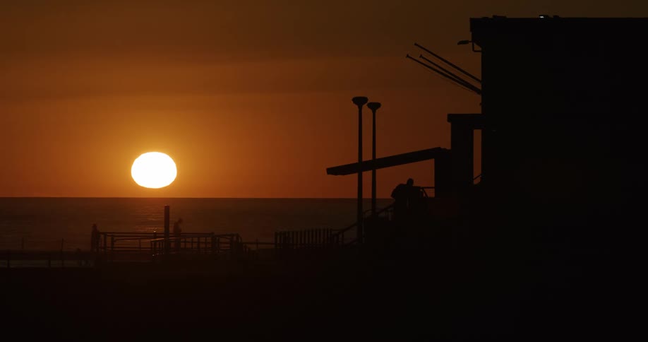 The sun sets over the ocean creating a serene scene with silhouettes of a person walking towards a house