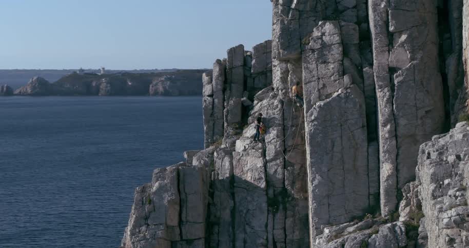 Two climbers ascend a steep rocky cliff beside a calm sea