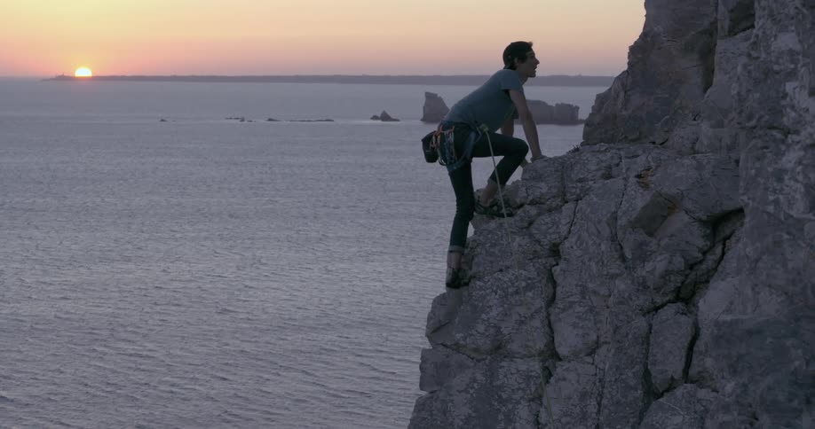A man with a backwards cap is climbing a mountain of rocks and sliding a rope down to an ocean with islands of rocks