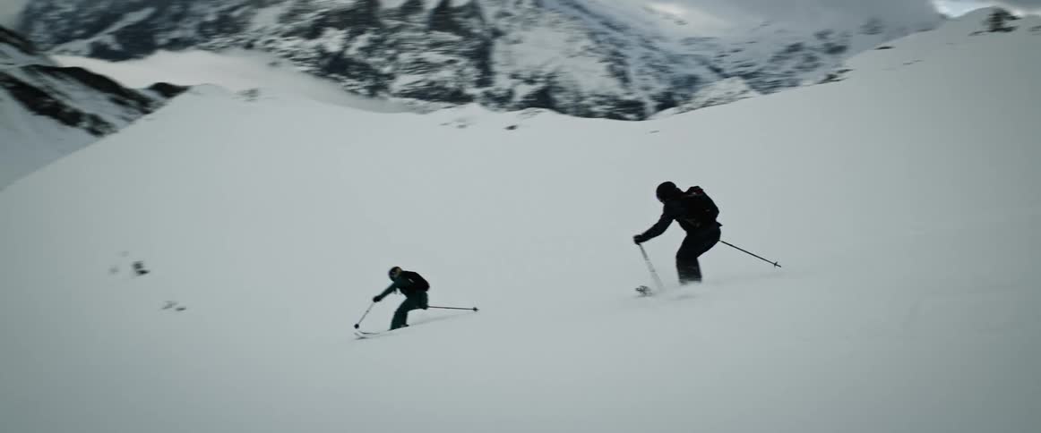 Two people skiing down a snow mountain and in front there are other stone mountains covered in snow