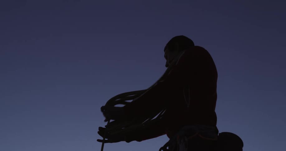 Silhouette of a man pulling a length of rope and arranging it while standing outdoors against a darkened sky