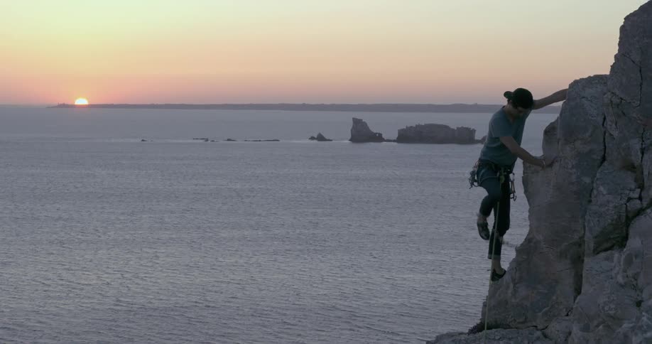 A man is climbing a mountain of rocks using a gear rope and below is an ocean and the sun setting on the horizon