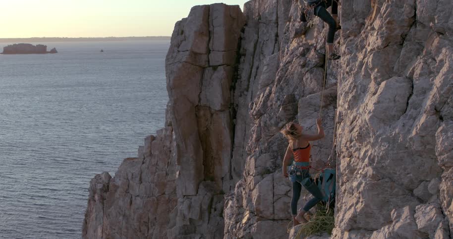 Woman on a cliff helping her colleague who is climbing the rocky wall of the coast by the sea