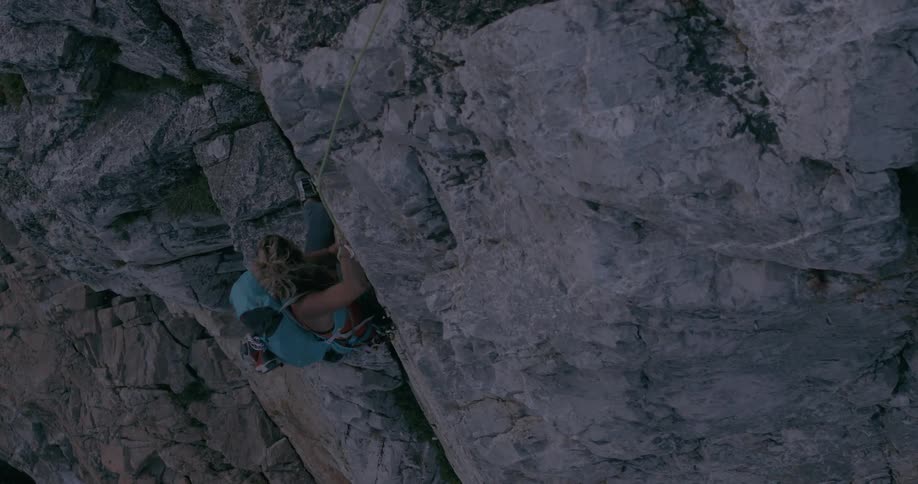 A blonde woman is climbing a rock using a rope and a blue backpack