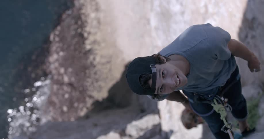 A man is hanging from a mountain of rocks using a rope and is looking up while smiling below him there is water moving