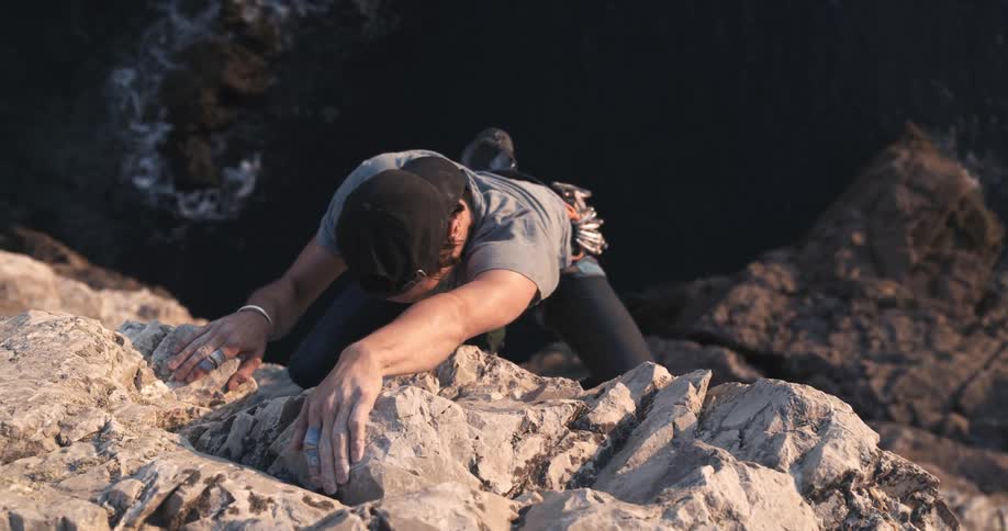 A young boy with a backwards cap is climbing up a mountain of rocks and down to the sea