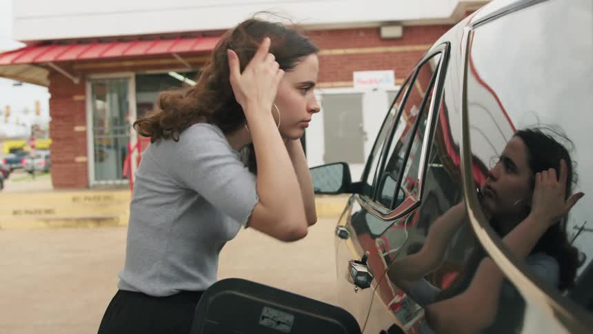 A young woman primps her hair while filling her car with gas