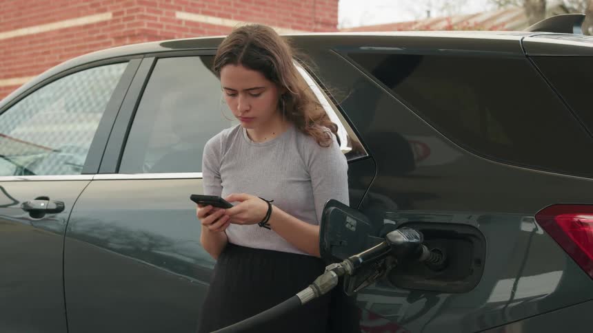 Woman leaning against her car playing on her cell phone waiting while her car is being filled with fuel