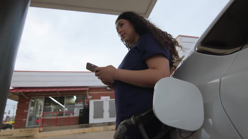 Young woman using her cell phone while her car is being filled up at a gas station