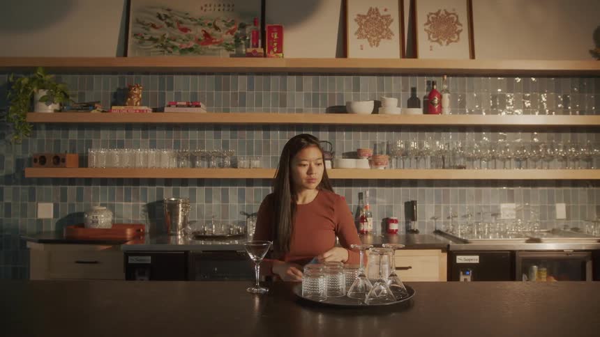Young woman at a counter drying crystal cocktail glasses and then placing them on a wooden shelf