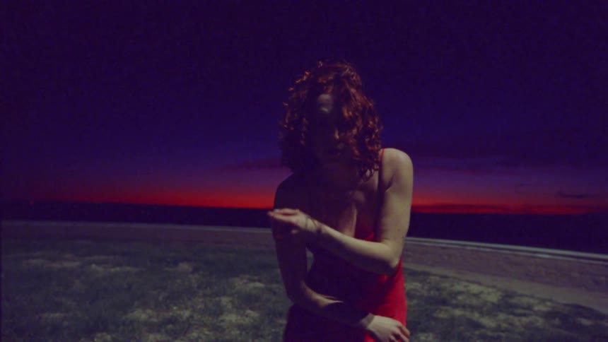 Woman with curly red hair and a red dress dancing in an open environment on a night with a red sky on the horizon
