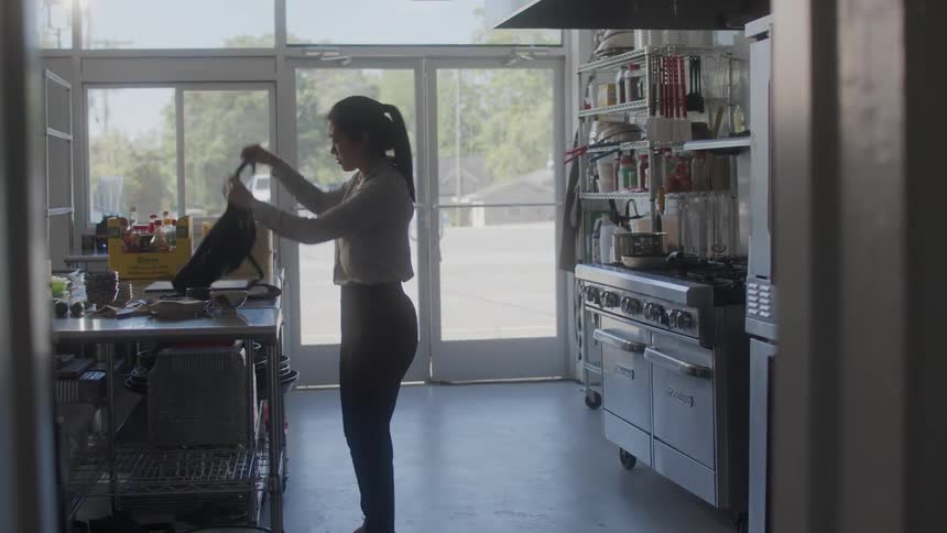 Asian female chef walks into restaurant kitchen and puts on apron while traffic passes on nearby street