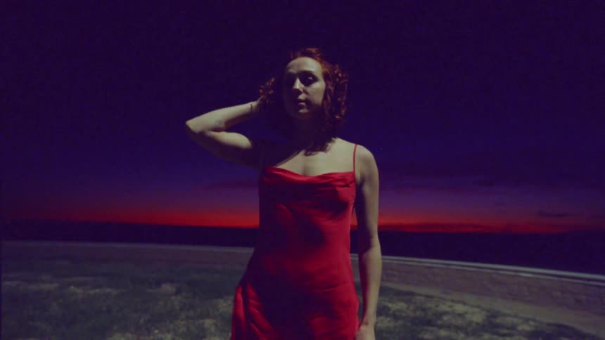 Woman running her hand through her curly red hair while wearing a red dress outdoors at night with the red sky