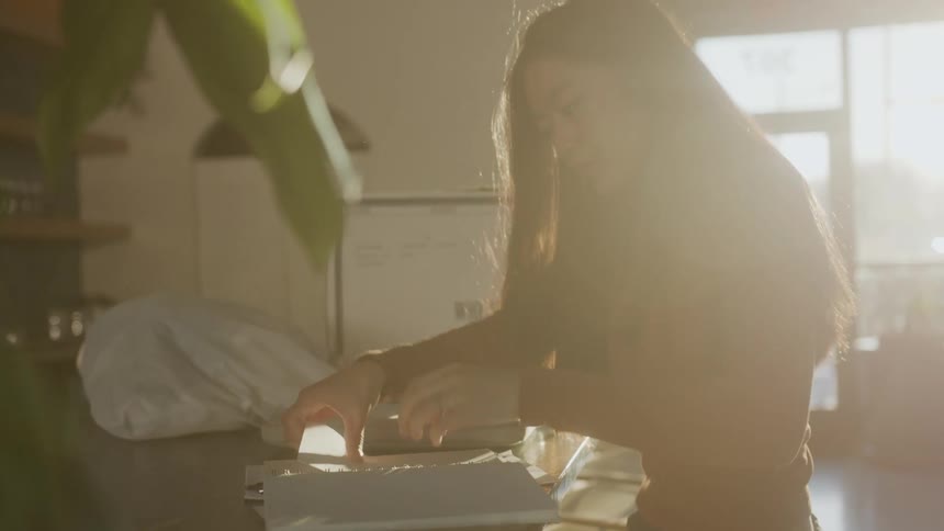 Woman carries in a box of alcohol with a notebook and laptop on top of it and sets it on the counter before opening the notebook and laptop to work