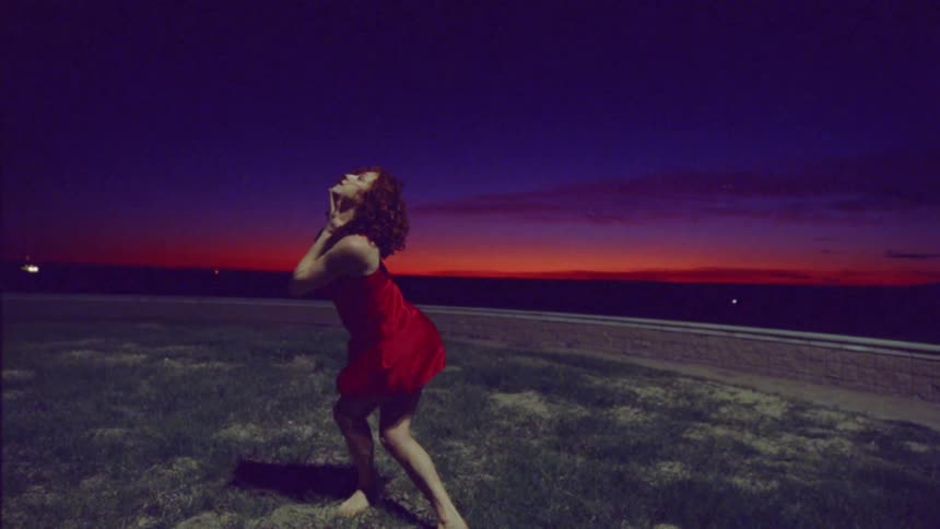 Woman dancing in a conical shape in a red dress on a lawn on a night with a reddish sky