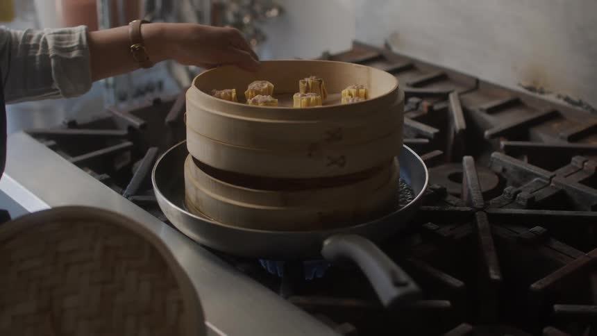 Frying pan with water on a stove and a womans hand placing a wooden bowl with asian cuisine dumplings to be cooked
