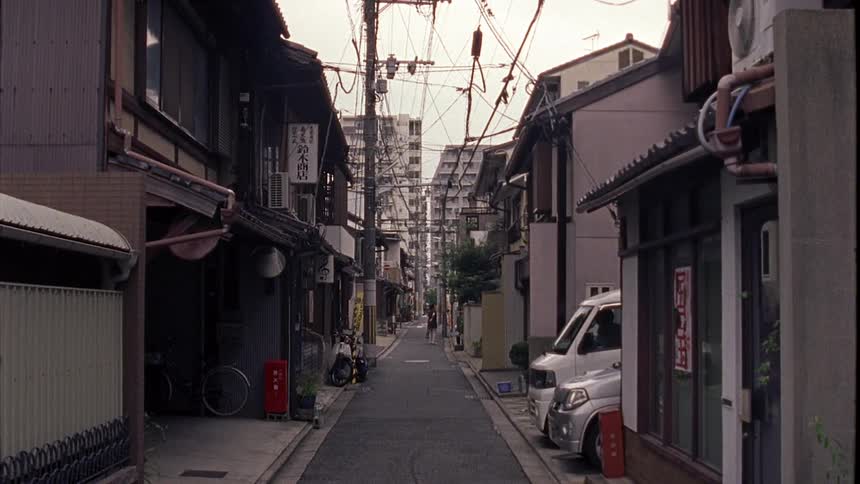 A narrow street with many wires and cables above and only a woman walking at the end