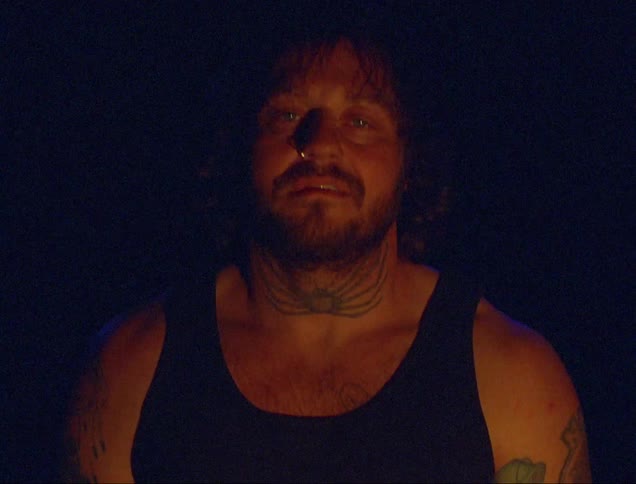 A man with neck tattoo in a tank top with people running behind him
