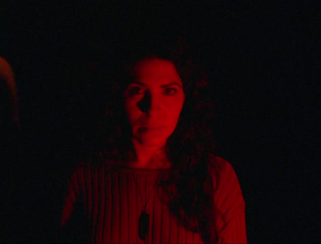 Portrait of a woman in a dark environment with low lighting and green and red lights passing over her face while people pass around her