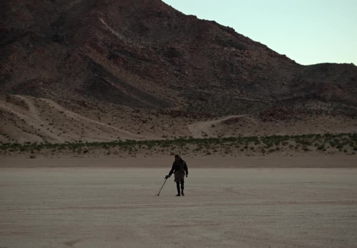 Man gently swings a metal detector back and forth over the sandy ground in front of a mountain