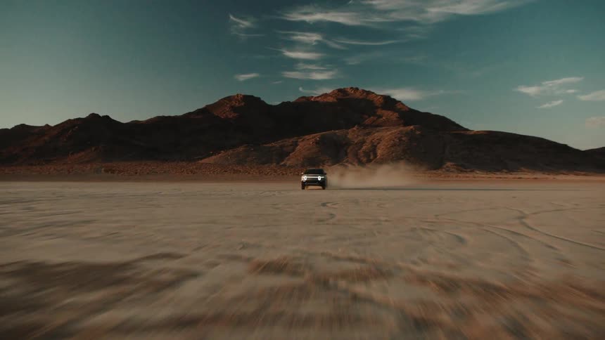 Truck speeds across the empty desert in front of a mountain on a sunny day