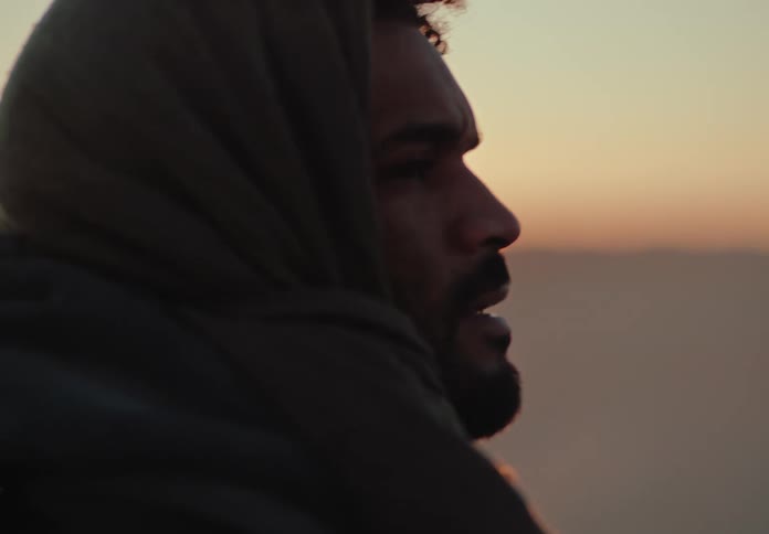 Side portrait of a man removing the scarf that is hanging over his mouth while standing in a natural environment with the sun on the horizon