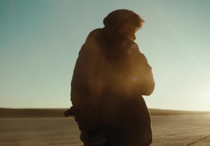 A man walking in the desert and running a metal detector across the ground in front of him removes the cloth protecting his mouth