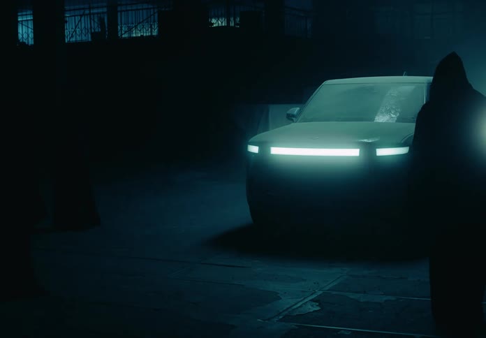 A hooded figure observes a pick up truck in a garage at night when its headlights turn on