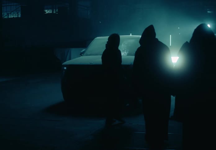 Hooded figures surround a pick up truck in a garage at night as a light moves around the truck