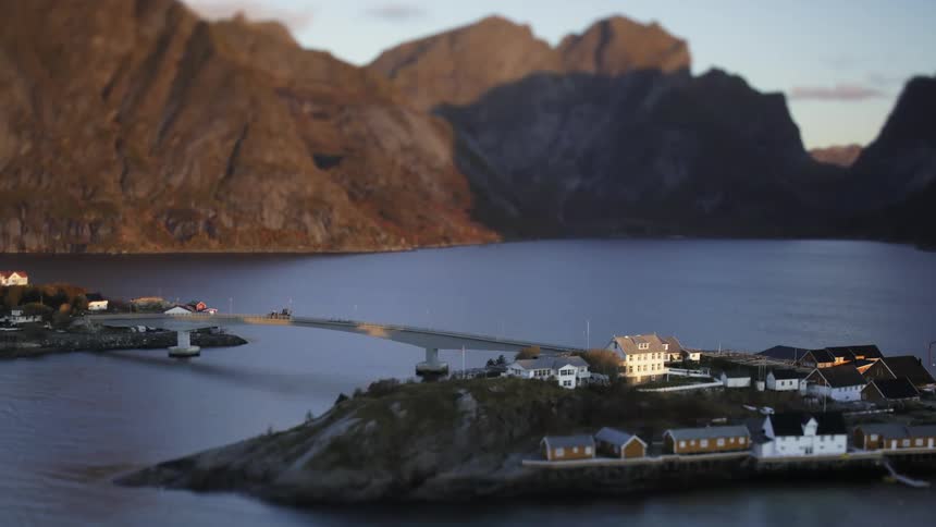 A car passing over a bridge that connects two pieces of land with many houses and around a lake with lots of water and behind it there are huge mountains