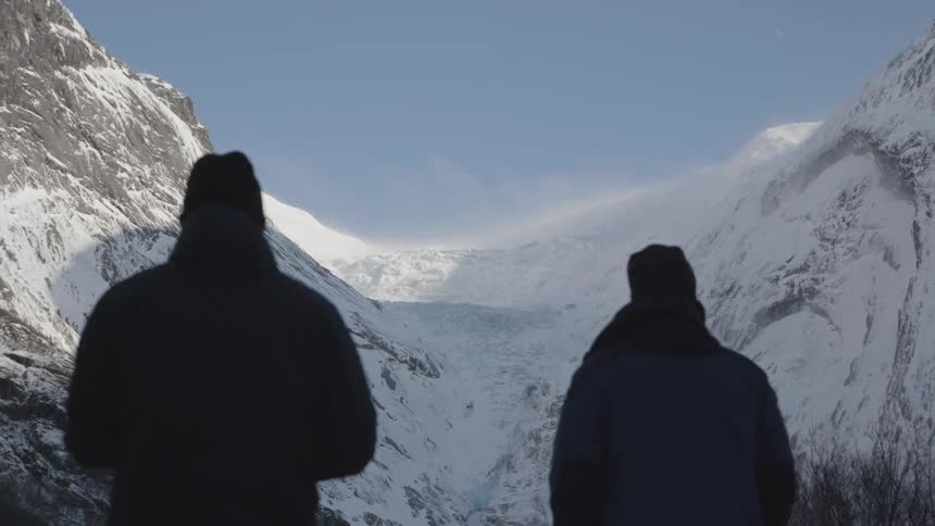View of two people from behind wearing headdresses while standing in a natural environment surrounded by snowcapped mountains on a cloudless blue sky day