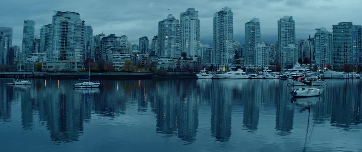 A cityscape with tall buildings reflected in calm waters with moored boats