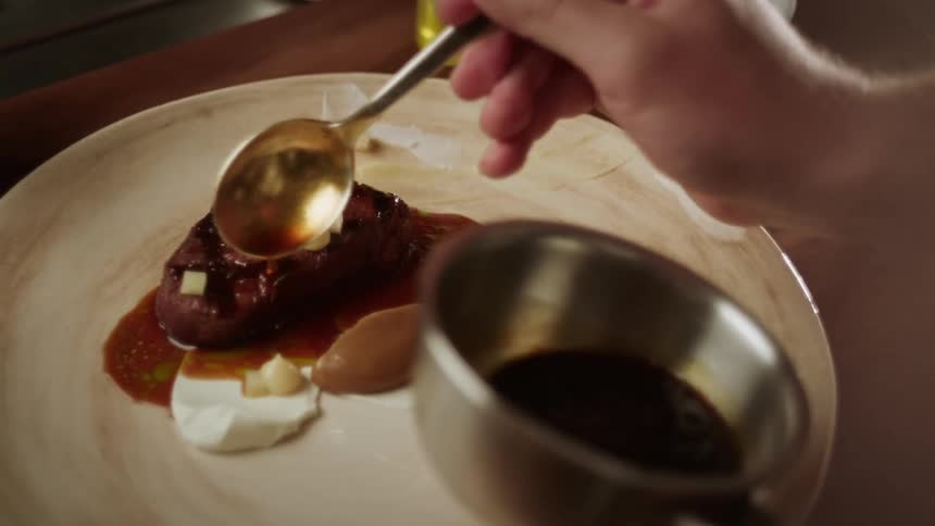 A persons hand pouring a sauce onto a plate with a piece of meat