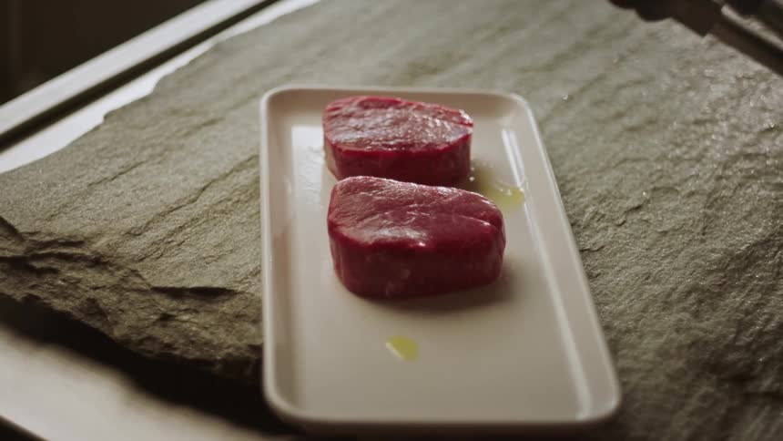 Tongs held over a small ceramic tray on a slap of stone with two chunks of meat