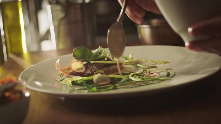 A persons hand putting oil from a white bowl onto a plate with vegetables