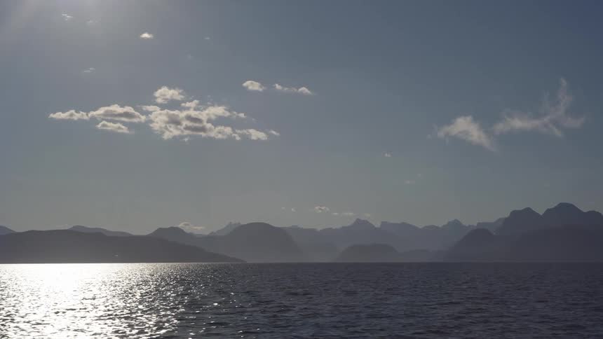 Sea landscape with sun reflection and mountain ranges on the horizon on a blue sky day with few clouds