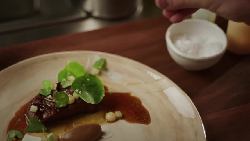 A mans hand is shown sprinkling salt onto a plate of steak and greens