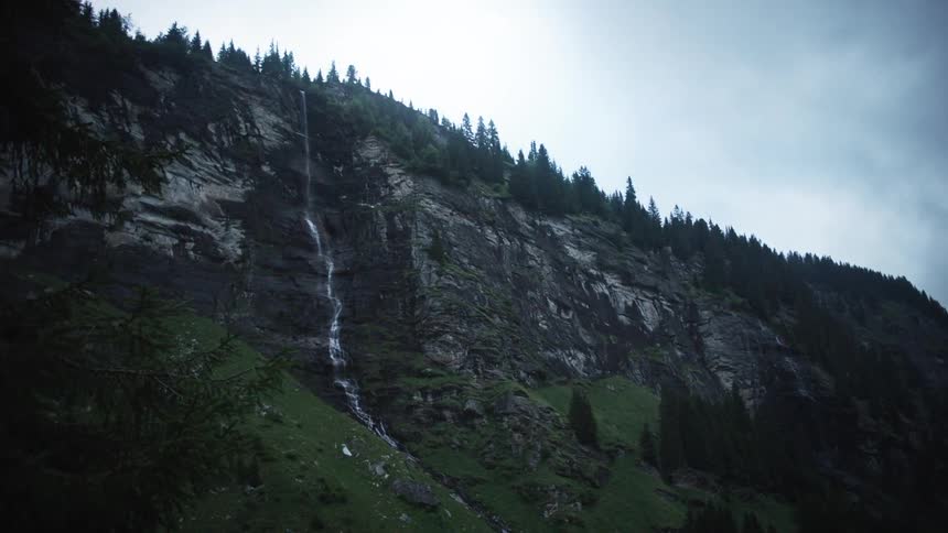 Mine from a waterfall on a rocky mountain wall with vegetation and conifers around on a cloudy day