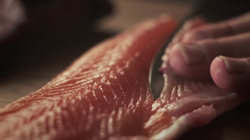 A persons fingers resting on a salmon and carefully cutting a piece of fresh salmon with a knife