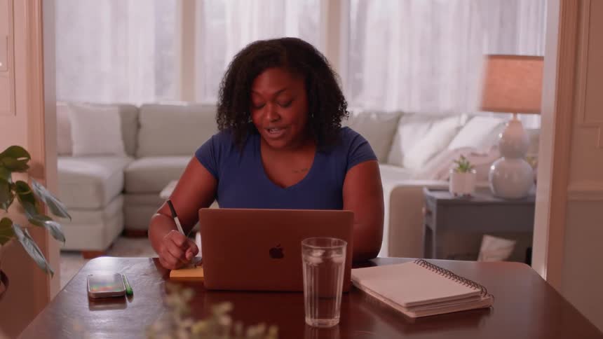 A woman is seated at a wooden table working on a laptop with a notebook