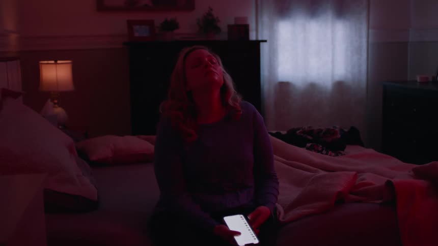 Woman sitting on the bed using her cell phone and then placing her hand on her face with a tired and discouraged facial expression
