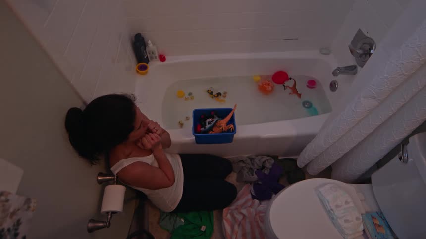 Woman inside a bathroom next to a bathtub putting her hand to her face with a discouraged facial expression and then picking up toys that are in the bathtub water