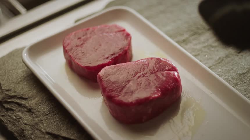 Two pieces of raw meat in an oil with a silicone spatula moving alongside