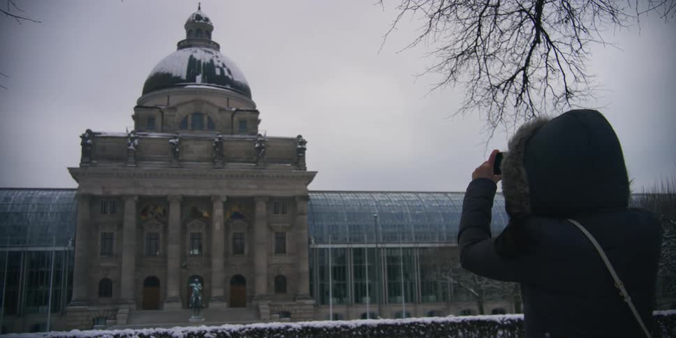 View of a woman in a hooded jacket with her smartphone pointed at the hermitage museum on a cloudy snowy day in winter